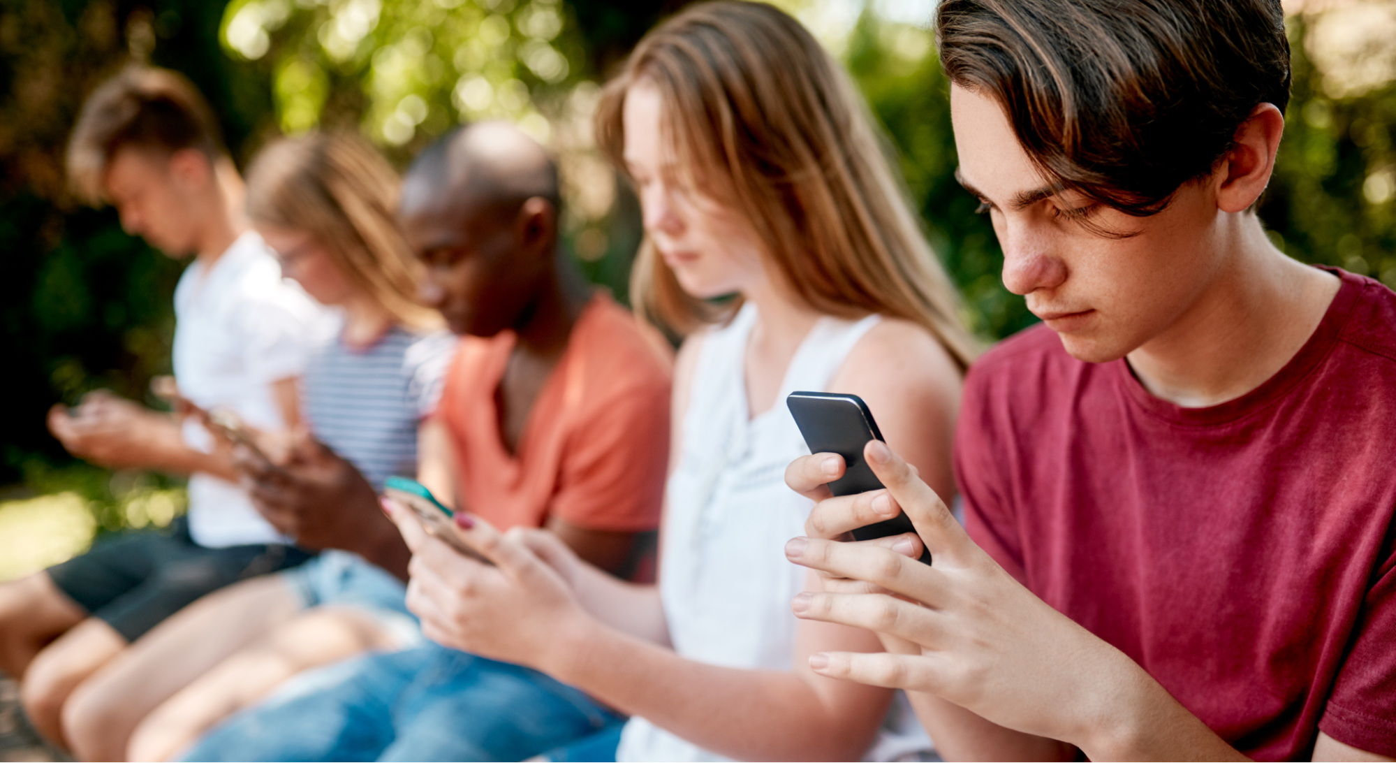 Five teenagers sitting in a row outdoors, each looking down at a smartphone