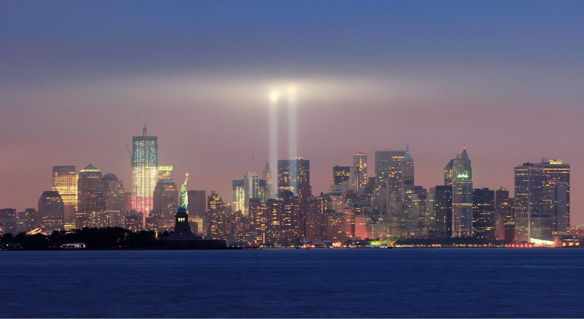 The Tribute in Light memorial rising above the Lower Manhattan skyline and the Statue of Liberty at dusk